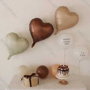 Peut inclure: Un gâteau au chocolat avec un glaçage blanc et des cerises rouges sur le dessus, entouré de ballons. Les ballons sont en forme de cœur et dans des tons de brun, beige et vert. Deux ballons blancs ont le texte "Happy Every Day" et "To You" imprimé dessus.