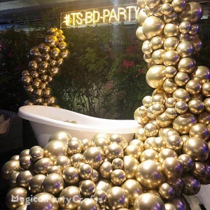 May include: A white bathtub with a gold balloon arch behind it. The balloons are arranged in a cascading waterfall design. The balloons are all the same size and are a shiny gold color. The text "#ITS-BD-PARTY" is visible in the background.