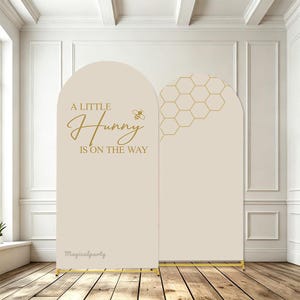 May include: Two arched backdrop panels in a light beige colour. The left panel features the gold text A LITTLE Hunny IS ON THE WAY with a small bee graphic. The right panel displays a gold honeycomb pattern. Both panels have gold metal bases.