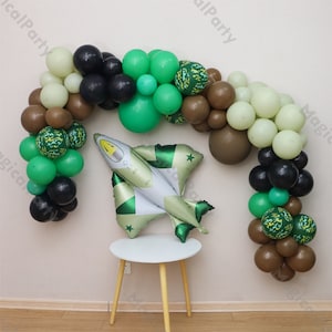 Pode incluir: Um arco de balões verde, marrom e preto com um padrão de camuflagem. Um balão de avião prateado está no centro do arco. O arco está em uma parede branca.