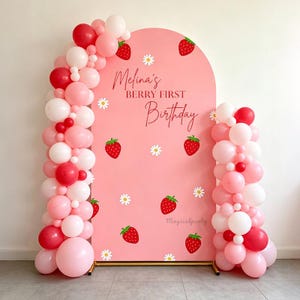 May include: A pink arched backdrop featuring strawberry and daisy illustrations with the text Melina's BERRY FIRST Birthday. The display is framed by a balloon garland in shades of red, light pink, and white, supported by a gold metal stand.