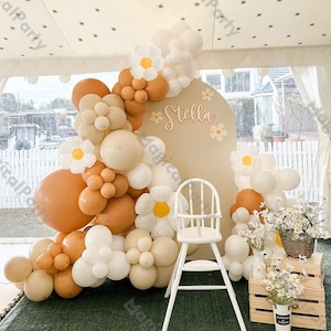 May include: A white high chair with a tan and white balloon arch decorated with white paper flowers. The balloons are arranged in a half-circle shape and the arch is decorated with the name "Stella".
