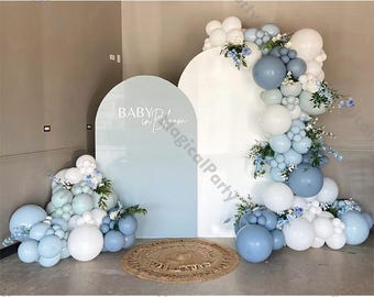 May include: A blue and white balloon arch with a white backdrop and a blue arch with the text "Baby in Bloom" in white. The balloons are arranged in a cascading design with blue and white balloons interspersed with greenery.