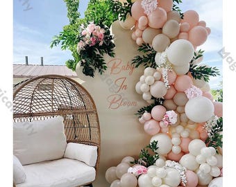 May include: A decorative backdrop for a baby shower, featuring a beige sign with the text "Baby In Bloom". The sign is adorned with pink and white balloons, greenery, and floral arrangements. A wicker chair with a white cushion is in the foreground.