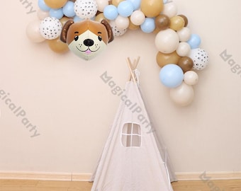 Puede incluir: Un globo de perro marrón y blanco con una guirnalda de globos azules y blancos. La guirnalda está colgada en una pared blanca con una tienda de campaña tipi beige en primer plano.