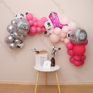 May include: A pink, silver and black balloon arch with a cow print balloon, a disco ball balloon and a pink high heel balloon. The arch is decorated with white cotton stems and a small white table with a brown paper bag and a picture frame.