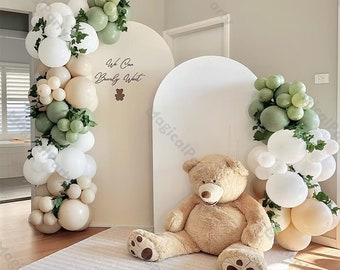 Könnte beinhalten: Ein weißer Torbogen, der mit grünen und cremefarbenen Ballons, Grünpflanzen und dem Text "We Can Barely Wait" dekoriert ist. Ein großer brauner Teddybär sitzt vor dem Torbogen.