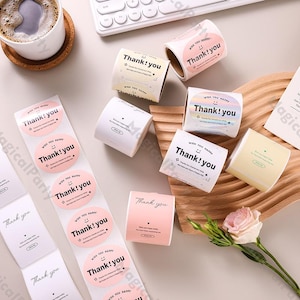 May include: A selection of "Thank you" stickers and labels in different colours and shapes. The stickers, some round and some rectangular, feature a smiling face graphic. Some are on rolls, others in sheets. A cup of coffee and a keyboard are in the background.