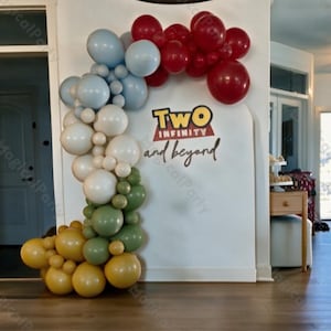 May include: A balloon arch featuring a color gradient of red, light blue, white, sage green, and mustard yellow balloons. The arch is positioned next to a white wall with the text "TWO INFINITY and beyond" printed in black, red, and yellow.