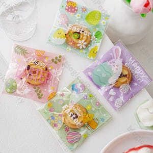May include: Four clear plastic bags with colorful Easter-themed designs. Each bag features a different design with a bunny, eggs, and flowers. The bags are filled with small cookies that say "Happy Easter".