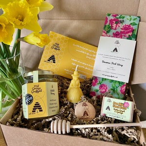 Puede incluir: Una caja de regalo llena de una variedad de artículos, que incluyen un tarro de miel, una envoltura de alimentos de cera de abejas, una vela, un cucharón de miel de madera, un pequeño adorno de colmena de madera y una bolsa de semillas de mezcla de abejas. La caja está forrada con virutas de papel marrón y decorada con narcisos amarillos.