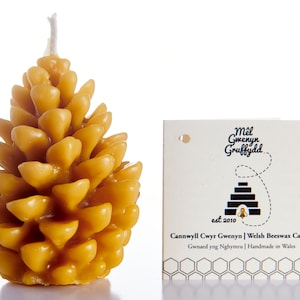 Könnte beinhalten: Eine gelbe Bienenwachskerze in Form einer Kiefernzapfen. Die Kerze ist handgefertigt in Wales und trägt den Text "Mêl Gmenyn Gruffydd est 2010 Cannwyll Cwyr Gwenyn | Welsh Beeswax Candle Gwnaed yng Nghymru | Handmade in Wales".