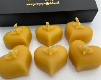 May include: Six heart-shaped, yellow beeswax candles with wicks, arranged in front of a black box. The box has gold text and a bee logo. The candles are on a white surface.