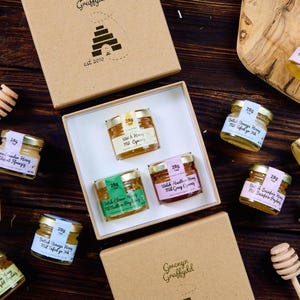 Op de afbeelding: Een verzameling kleine glazen potjes gevuld met honing, elk gelabeld met verschillende soorten. De potjes zijn gerangschikt in een geschenkdoos en op een houten oppervlak, samen met een houten honingdipper. De verpakking is bruin met een logo.