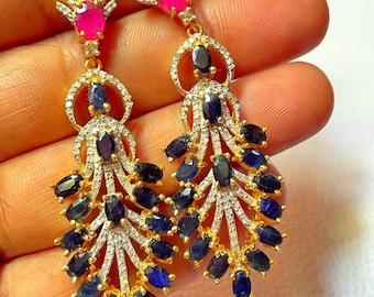 May include: A pair of gold earrings with a dangling design. The earrings feature a large oval-shaped blue sapphire surrounded by smaller sapphires and diamonds. The top of the earrings feature a pink ruby.