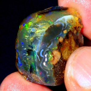 May include: A raw, polished opal gemstone featuring a translucent, glassy surface with vibrant flashes of green, blue, and red play-of-color. The stone displays a natural, irregular shape with dark, earthy brown inclusions and a smooth, reflective texture.