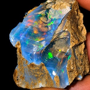 May include: A raw, unpolished opal specimen featuring a translucent, milky blue base with vibrant flashes of green, orange, and yellow play-of-color. The stone has a jagged, irregular shape with rough, earthy brown rock matrix along the edges.