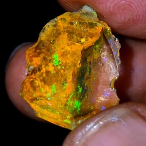 Puede incluir: Una piedra preciosa de ópalo bruto, translúcida, de color amarillo con iridiscencia verde y púrpura.