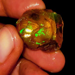 Op de afbeelding: Een ruwe, ongepolijste opaal edelsteen met een doorschijnende amberkleurige basis. De steen vertoont levendige flitsen van neongroen en oranje op het onregelmatige, getextureerde oppervlak tegen een donkere, effen zwarte achtergrond.
