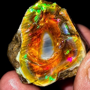 May include: A raw, unpolished opal gemstone featuring a vibrant play-of-color with intense flashes of neon green, fiery orange, and bright red. The center displays a smooth, translucent greyish-white core surrounded by textured, earthy rock.