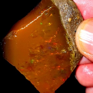 May include: A raw, translucent amber-colored opal gemstone featuring a rough, textured brown rock matrix along one edge. The polished section of the stone displays internal flashes of red, orange, and green fire against a warm, honey-toned base.