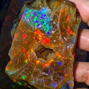 May include: A large, raw Welo opal gemstone weighing 649 Ct, featuring a translucent brown base with vibrant flashes of red, green, and blue play-of-color. The stone has a textured, cracked surface and a central cavity. Text reads 649 Ct and UNEARTHING THE POTENTIAL NATURAL WELO GEOLOGICAL WONDER.