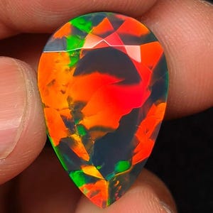 May include: A pear-shaped black opal gemstone featuring a dark base color with vibrant, intense flashes of neon orange and bright green play-of-color throughout the faceted surface, displaying a high-contrast pattern of light and dark areas.