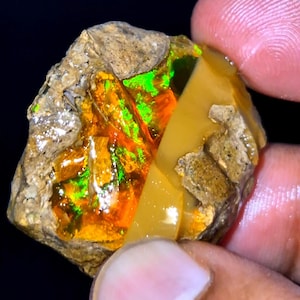 Könnte beinhalten: Ein roher, unpolierter Opal-Edelstein mit einer rauen, texturierten braunen Außenseite und einem lebendigen, polierten Innenbereich, der intensive Blitze in Neongrün und feurigem Orange auf einer glatten, durchscheinenden goldgelben Basis zeigt.
