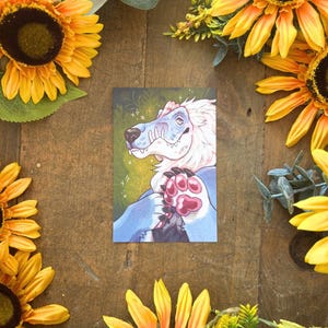 May include: A rectangular art print featuring a stylised illustration of a blue and white creature with a snout, sharp teeth, and pink paw pads. The creature is set against a dark green background with subtle floral line art and small star accents.