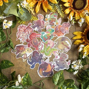 Può includere: Una collezione di adesivi colorati e stravaganti con vari personaggi di rettili e anfibi, tra cui tartarughe, serpenti e alligatori. Gli adesivi sono disposti su una superficie di legno, circondati da girasoli, verde e fiori bianchi.
