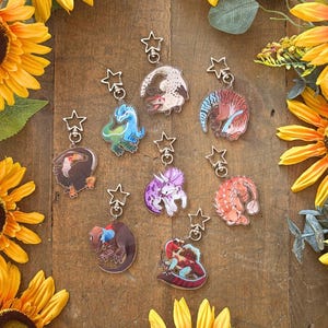 May include: Eight acrylic keychains featuring various dinosaur illustrations, each attached to a silver-coloured star-shaped clasp. The designs include a blue and green raptor, a purple triceratops, a red and blue creature, and other prehistoric animals.