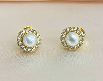 May include: A pair of gold stud earrings with a white pearl center and a clear crystal halo.