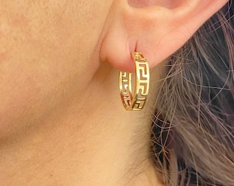 May include: Gold-coloured hoop earrings featuring a Greek key design. The earrings are worn on the ear and have a geometric pattern. The earrings are a bright gold colour and are a classic jewellery item.
