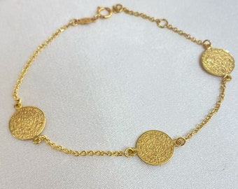 Puede incluir: Pulsera dorada con tres colgantes circulares texturizados, unidos por una fina cadena. La pulsera se presenta sobre una superficie blanca, resaltando su color dorado. La cadena mide aproximadamente 18 cm.