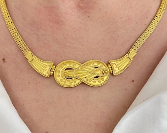 Könnte beinhalten: Eine goldene Halskette mit einem großen, kunstvollen Anhänger in Knotenform. Der Anhänger ist mit einem sich wiederholenden Muster aus kleinen Kreisen und Linien verziert.