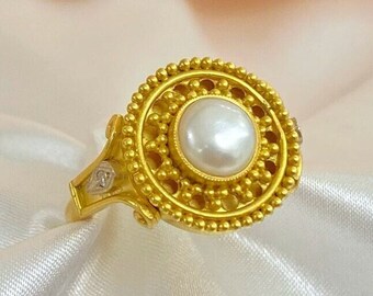May include: A gold ring featuring a large, round, white pearl at the center. The ring has a detailed, circular design around the pearl, with small, raised dots. The band has a decorative design with small, clear stones.