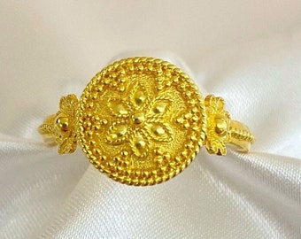 May include: A gold ring with a round, ornate design. The ring features a central flower motif surrounded by a beaded border.