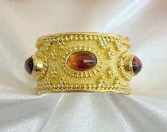 May include: A gold ring with a textured band and three oval-shaped gemstones. The center stone is brown, and the two side stones are a darker shade of red.