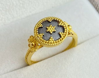 May include: A gold ring featuring a circular face with a dark gray, textured center and a small gold star-shaped detail. The ring band is decorated with a rope-like pattern and a small floral accent on one side, showcasing intricate metalwork.