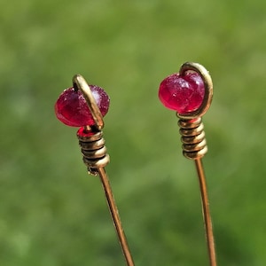 Könnte beinhalten: Nahaufnahme von zwei handgefertigten Haarnadeln. Jede Nadel hat eine tiefrote, facettierte Perle an der Spitze, die von einem goldfarbenen Draht umgeben ist. Die Nadeln haben ein Spiraldesign und einen schlanken, goldfarbenen Stiel. Der Hintergrund ist ein weiches Grün.