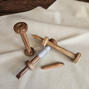 Könnte beinhalten: Drei Holzspulen mit natürlicher Maserung, eine davon mit hellgrauem Garn umwickelt. Die Spulen haben einen zylindrischen Schaft mit ausgestellten Enden und einer Spitze. Der Text www.lindwurm-couture.de ist unten aufgedruckt.