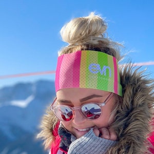Può includere: Una fascia colorata con strisce rosa, gialle e verdi è indossata. La fascia ha la scritta "evn" in bianco. La persona indossa occhiali da sole e una giacca rosa con cappuccio bordato di pelliccia. Lo sfondo mostra una catena montuosa e un cielo azzurro.