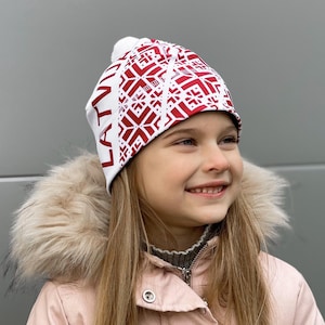 Puede incluir: Un gorro de punto blanco y rojo con un pompón en la parte superior. El gorro tiene un patrón letón y la palabra "LATVIA" escrita en el lateral.