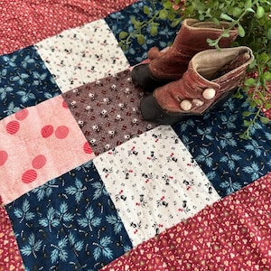 Könnte beinhalten: Eine rote, weiße und blaue Patchwork-Decke mit Blumenmuster. Zwei braune Lederstiefel mit weißen Knöpfen liegen auf der Decke.