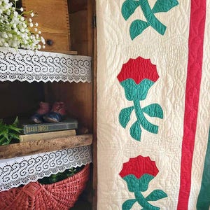May include: A white quilt with a red and green floral pattern. The quilt is hanging on a wooden cabinet with white lace trim on the shelves.