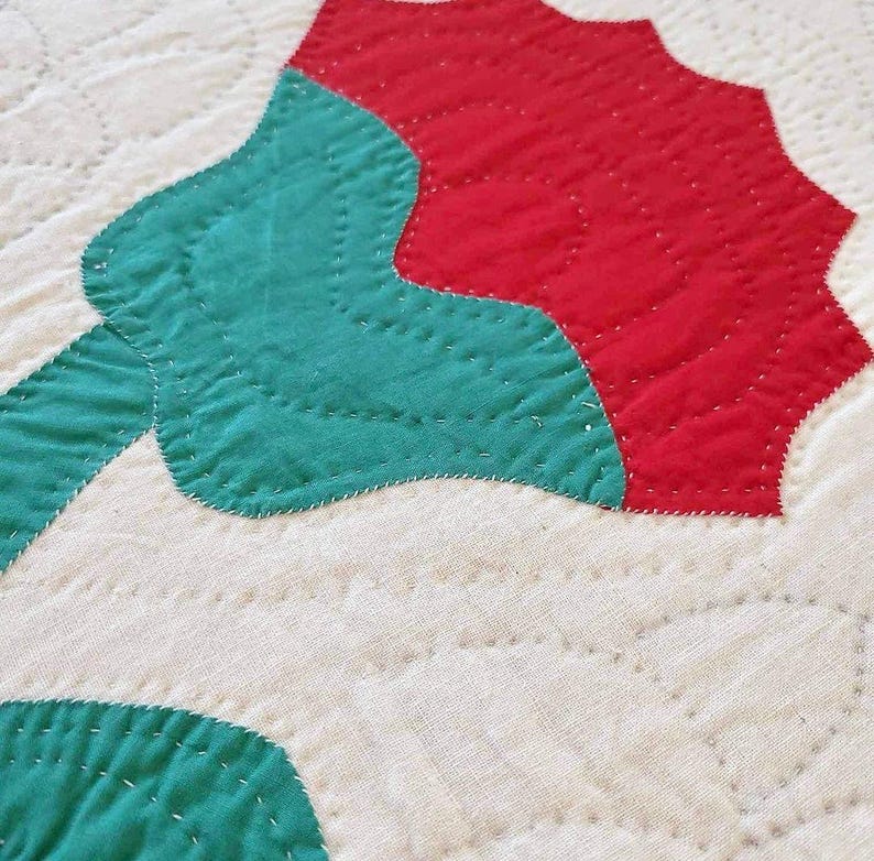 May include: A close-up of a red and green quilt block with a flower design. The quilt block is made of fabric squares sewn together with a white thread.