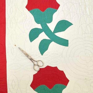 May include: A white quilt with red and green applique roses. The quilt is quilted with a circular pattern. A pair of silver scissors are lying on the quilt.