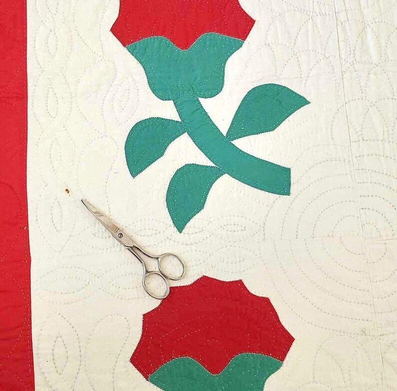 May include: A white quilt with red and green applique roses. The quilt is quilted with a circular pattern. A pair of silver scissors are lying on the quilt.