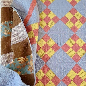 May include: A blue, yellow, and pink patchwork quilt with a diamond pattern. The quilt is folded in half and the edges are visible.