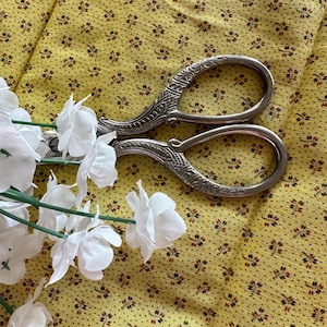 Könnte beinhalten: Eine silberne Schere mit verzierten Griffen liegt auf einem gelben Stoff mit kleinem Blumenmuster. Im Vordergrund befinden sich weiße Blumen.